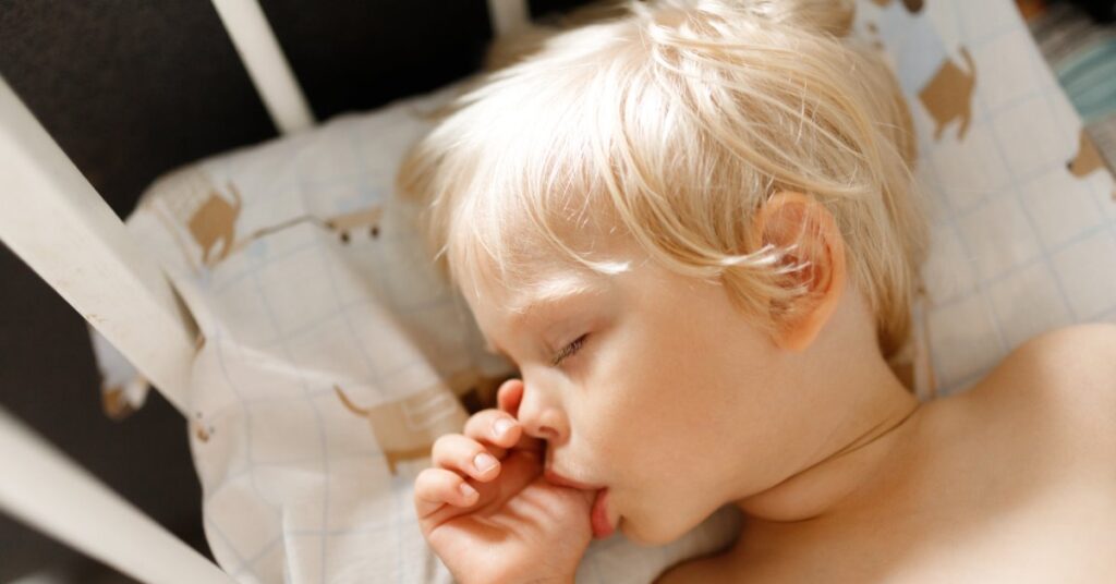A blond child sleeping in a white crib with a hand near the mouth on a patterned pillow in warm light.