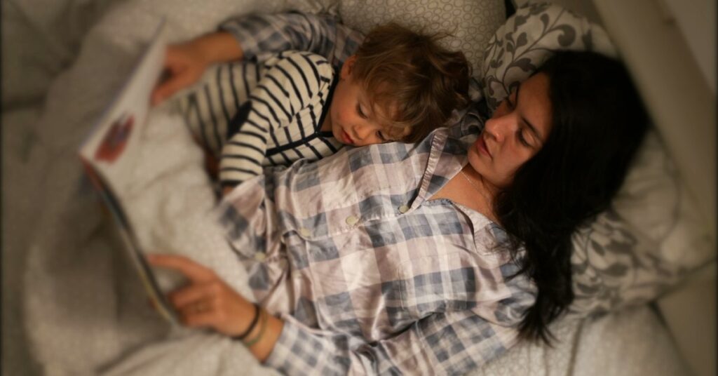 A young child lies curled beside a woman on a bed, and they read a book together. Both wear patterned sleepwear.